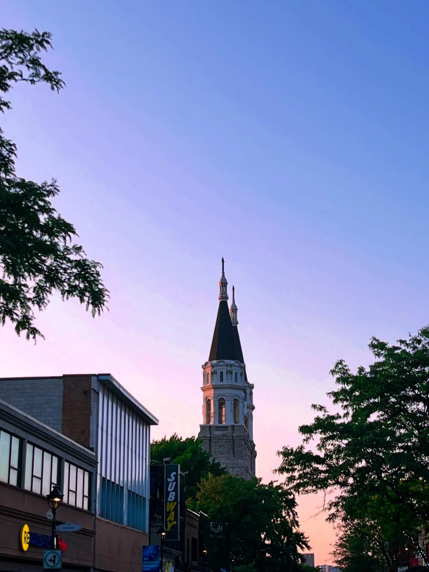 A church spire catching the last light at dusk, above the shop signs of a Montréal street