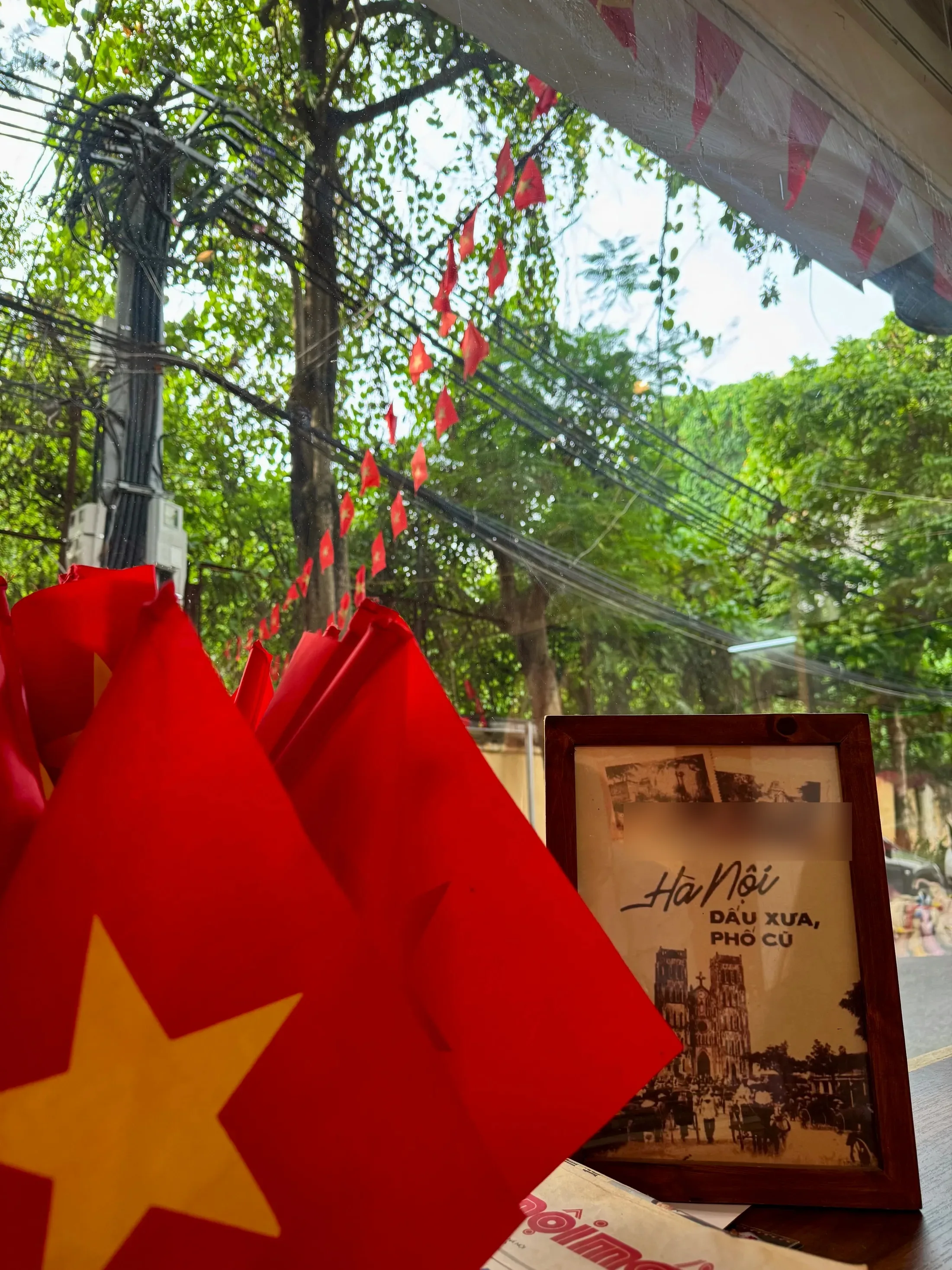 Small Vietnamese flags on a table by a window on a Hanoi street, beside a framed poster of St Joseph’s Cathedral