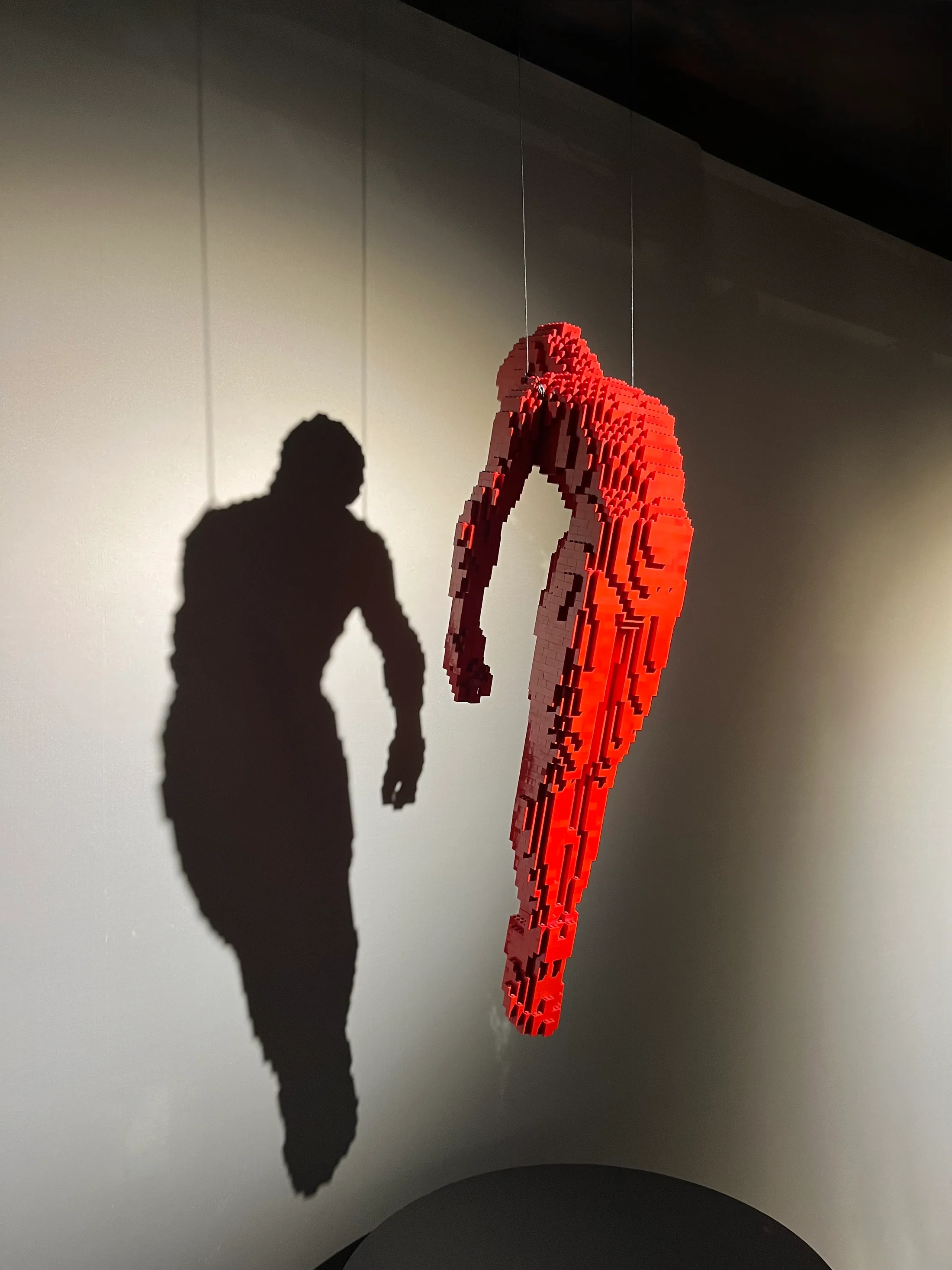 A red Lego figure suspended mid-air in a gallery, its shadow thrown large on the wall behind it