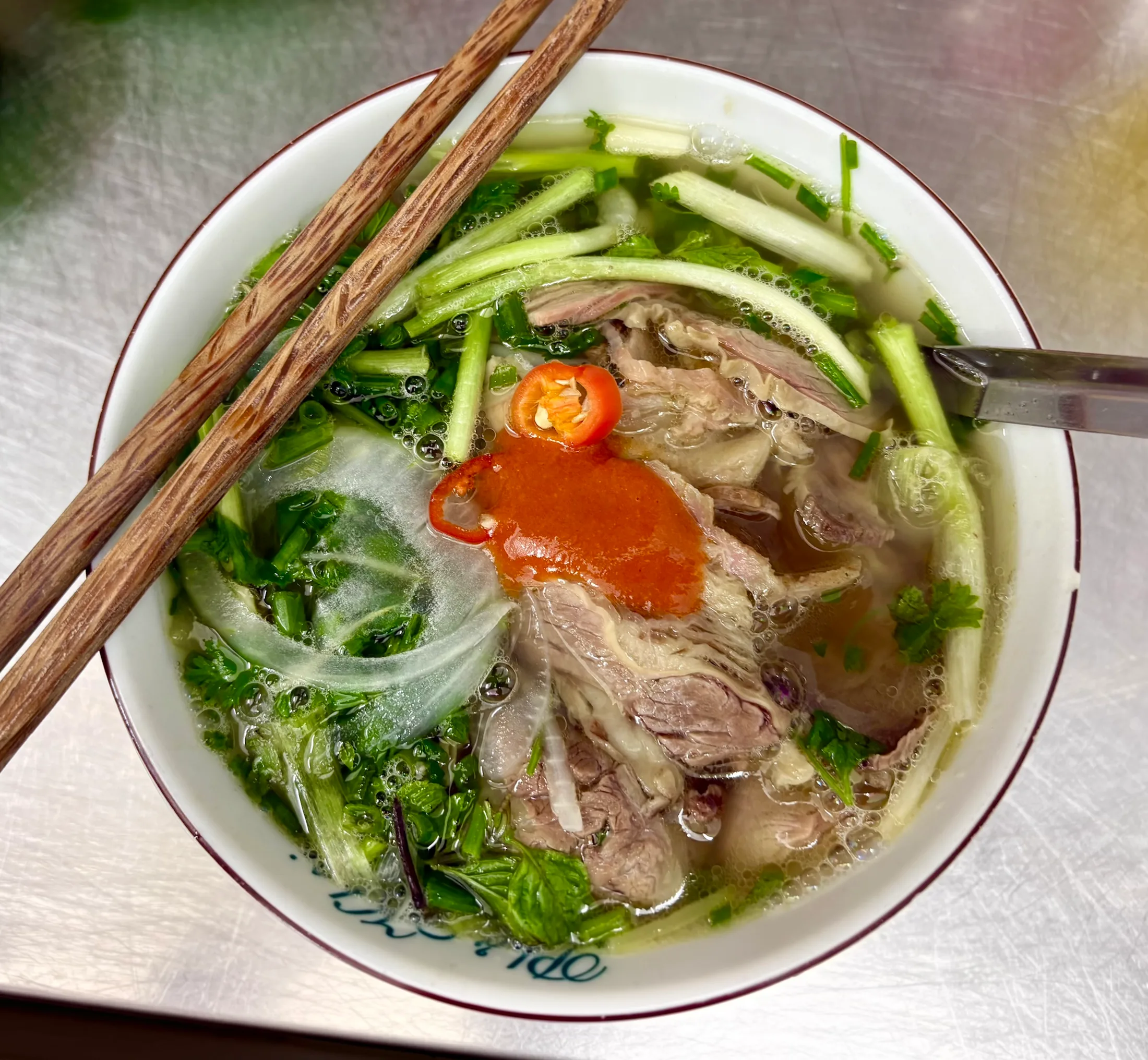 A bowl of phở from above, chopsticks across the rim, a spoonful of chilli sauce and a slice of red chilli on the beef and spring onions