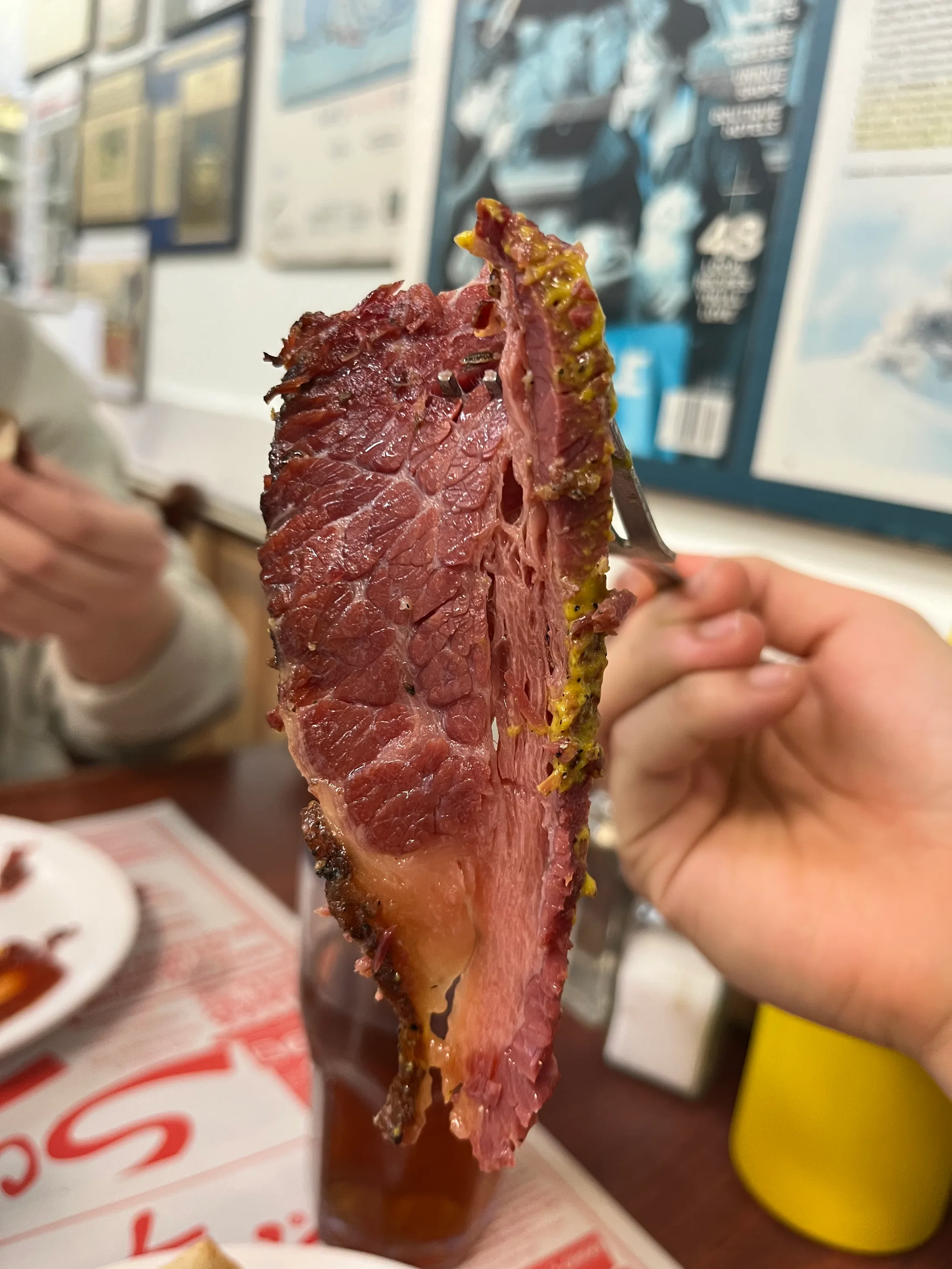 A hand holding up a slice of Montréal smoked meat, framed clippings on the wall behind